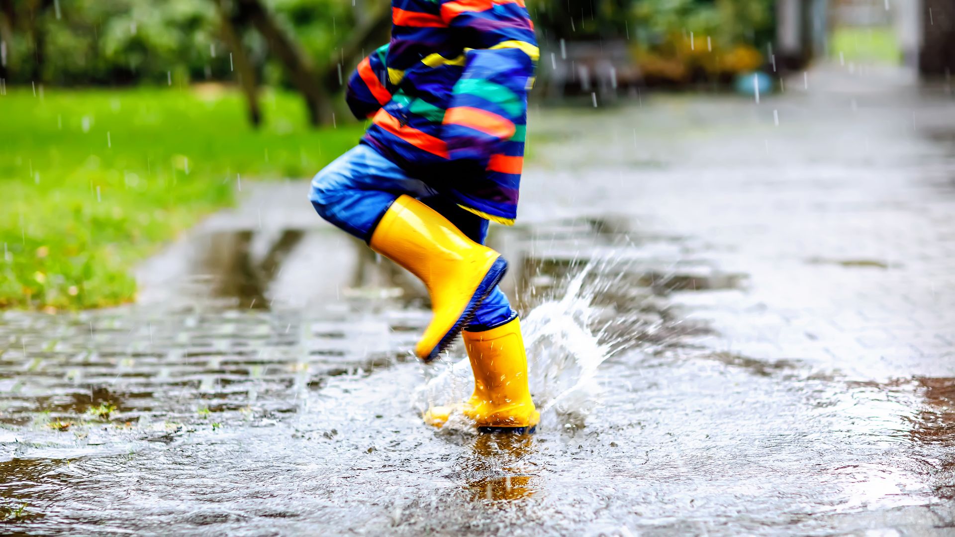 Ein Kind mit gelben Gummistiefeln und buntem Regenmantel springt in eine Pfütze.